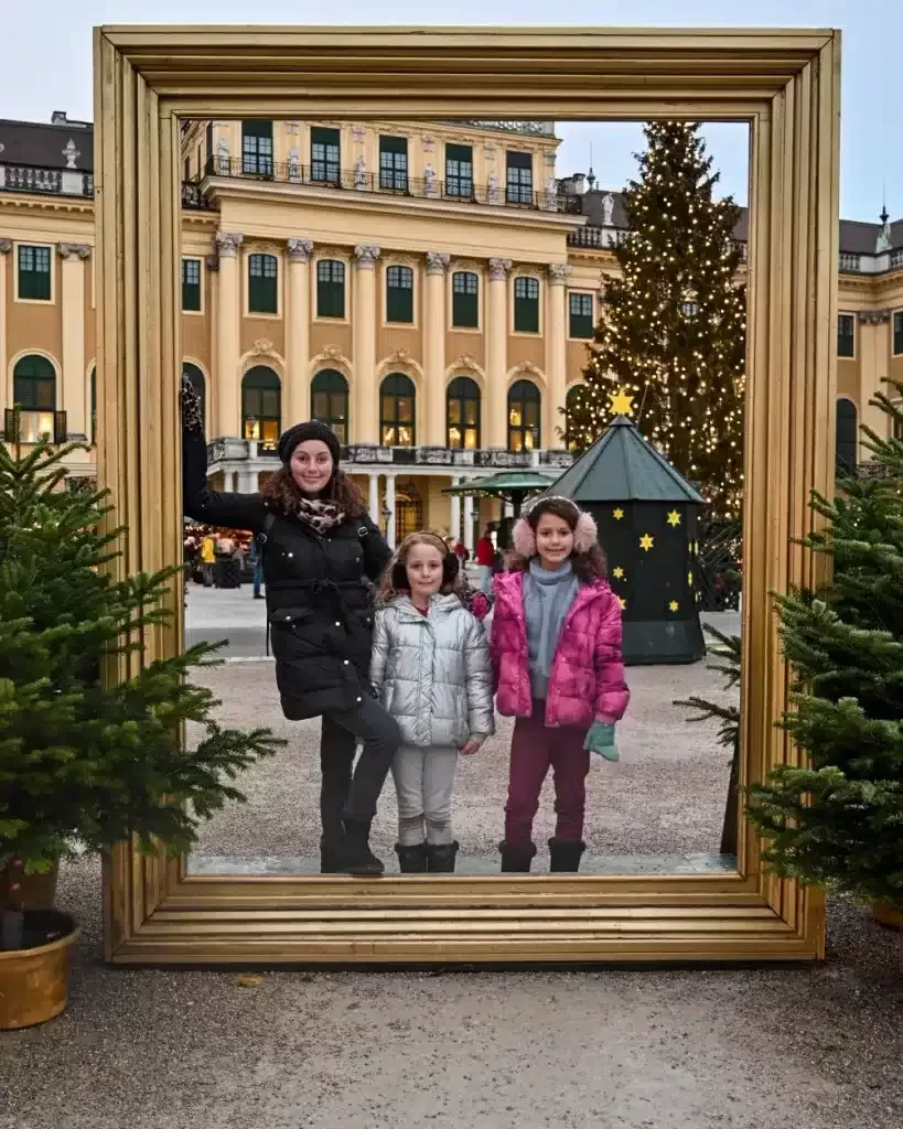 Mother and daughters standing in a giant golden frame in front of Schonbrunn Palace in Vienna during a Christmas Market