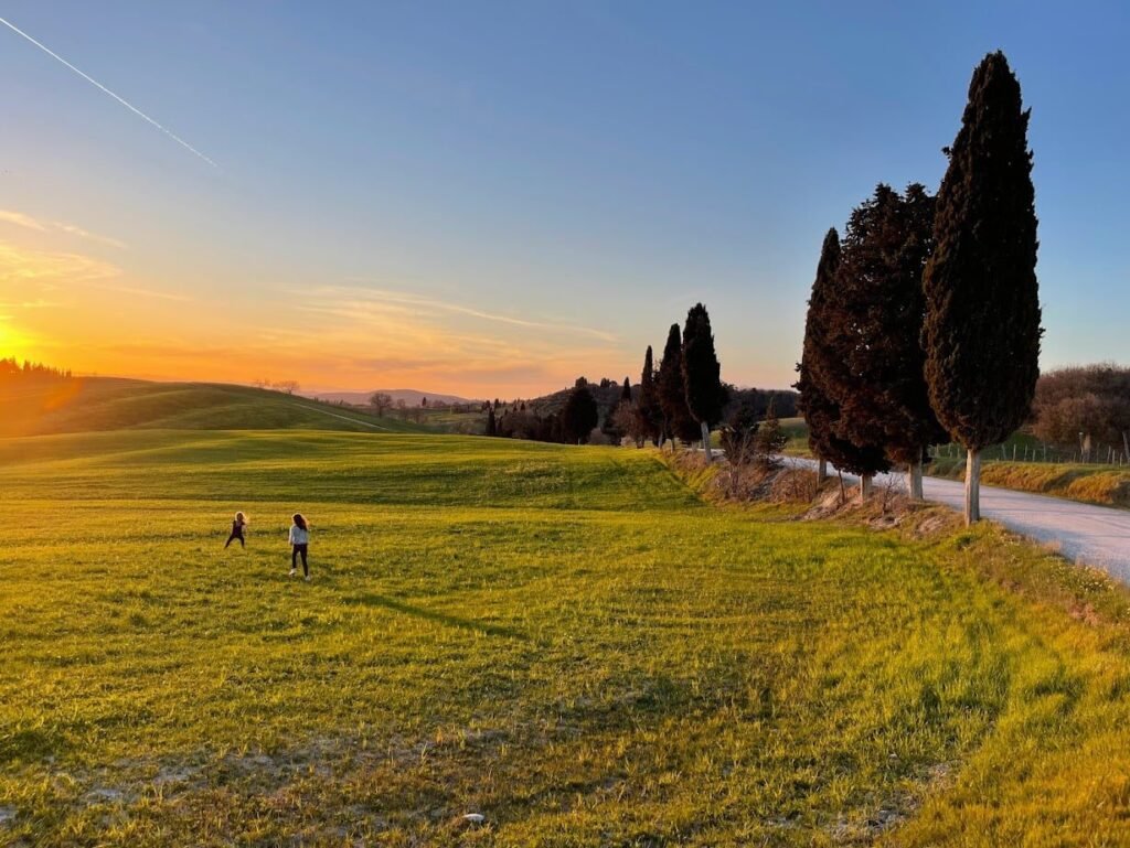 Epic views are one of the best parts of visiting Tuscany with Kids. Kids playing during a Tuscany family vacation in a field lined by cypress trees at sunset