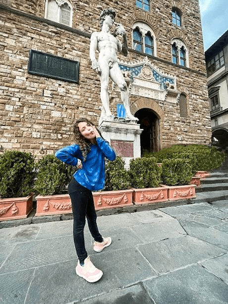 Enjoying art is a must when in Tuscany with kids. A little dark haired girl makes a silly pose in front of outdoor David statue in Florence