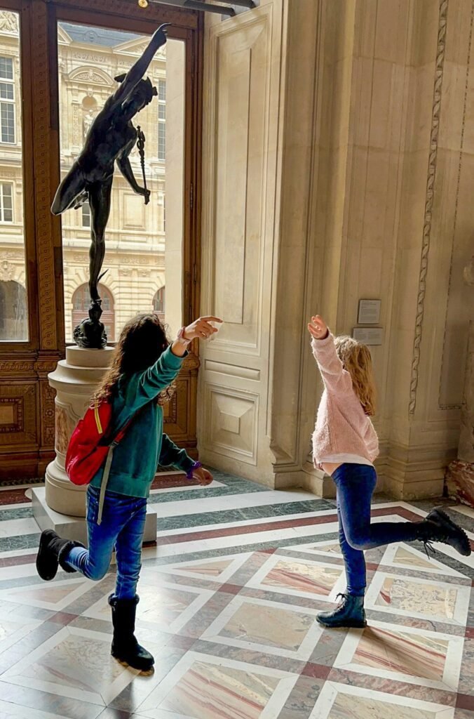 A stop at the louvre in Paris with kids as two little girls pose like a sculpture