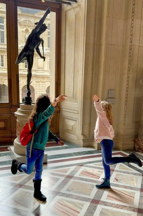 A stop at the louvre in Paris with kids as two little girls pose like a sculpture