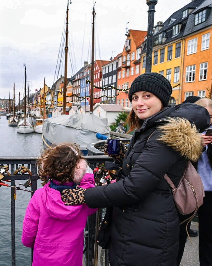 Woman looking over her shoulder and wearing a black parka in Nyhaven neighborhood in Copenhagen. She is standing with her daughter who is facing away from the camera towards the water.