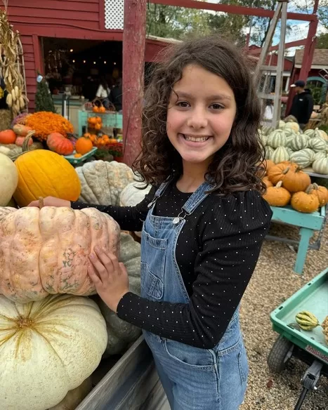 Little girl in overalls smiling in front of pumpkin stand at Depaul's Urban Market in Vienna, VA