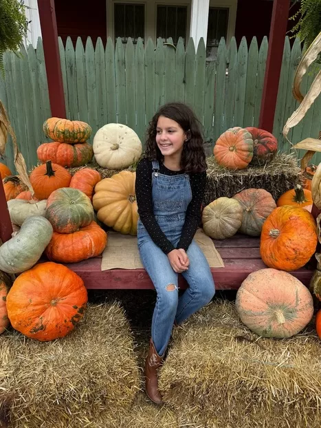 Little girl in overalls smiling in front of pumpkin stand at Depaul's Urban Market in Vienna, VA